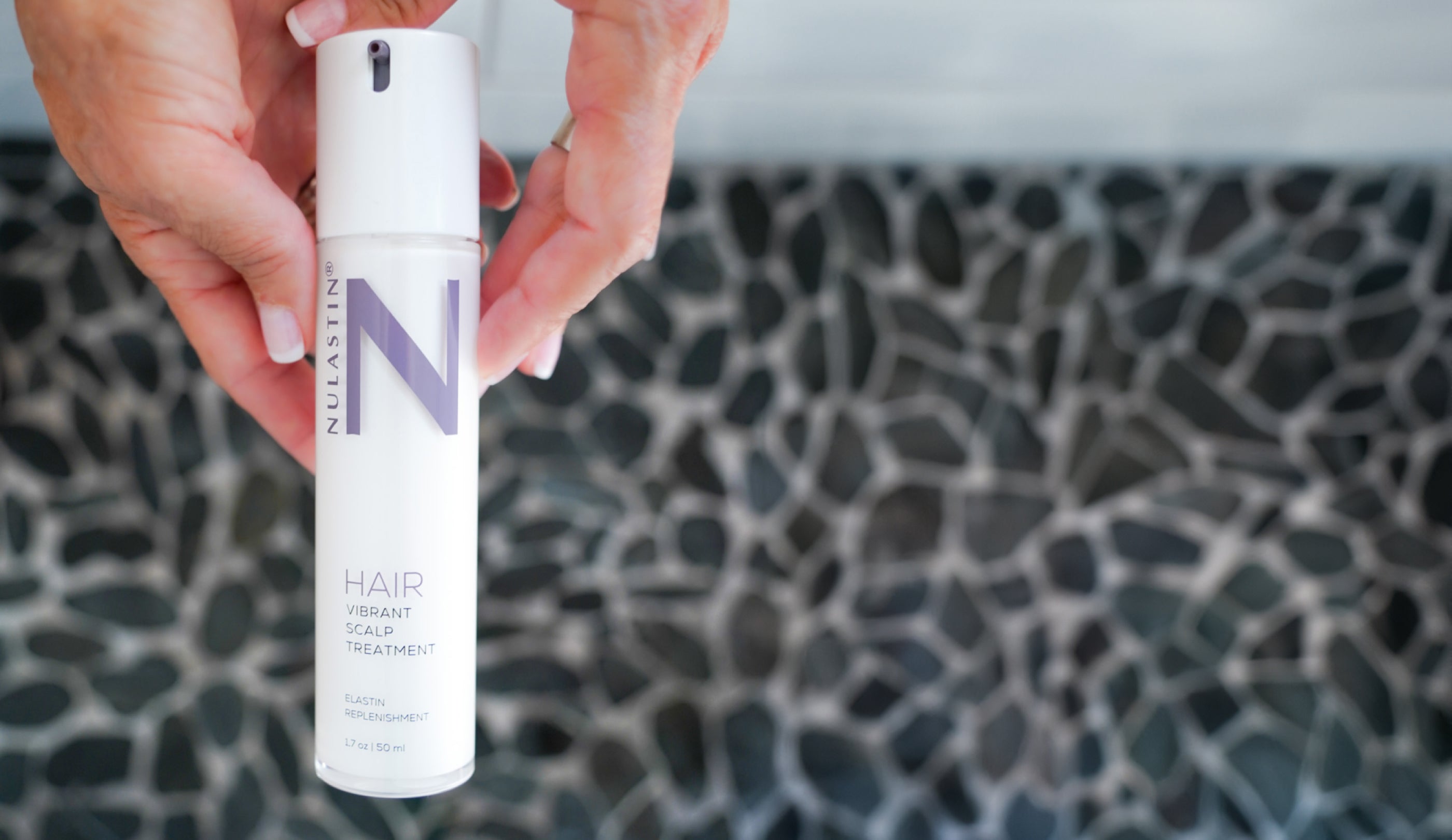 Manicured hands hold a bottle of NULASTIN HAIR Vibrant Scalp Treatment in a beautiful, spa-inspired bathroom.