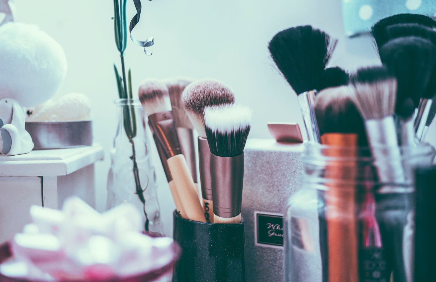 Assorted makeup brushes in jars and holders on a vanity counter with soft pastel lighting