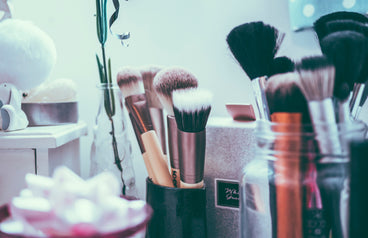 Assorted makeup brushes in jars and holders on a vanity counter with soft pastel lighting