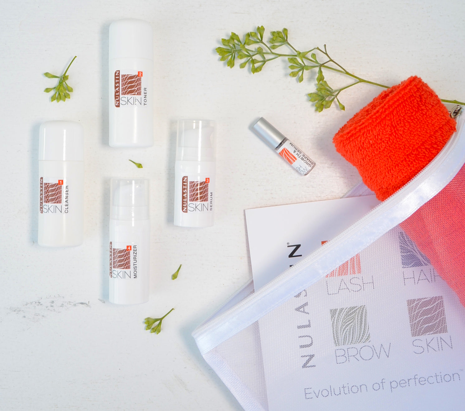 NULASTIN travel skincare flat lay with four travel-size bottles (cleanser, toner, serum, moisturizer), a mini treatment, orange washcloth, and a branded mesh travel pouch