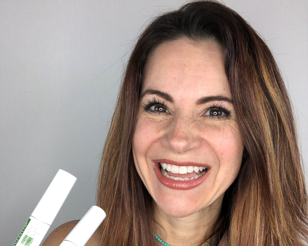 Woman smiling and holding two NULASTIN Lash and Brow Dual System tubes against a plain gray background