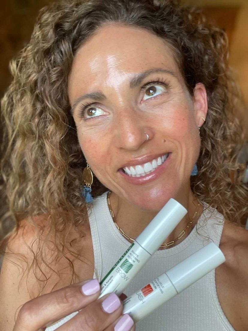 Smiling woman holding two NULASTIN lash and brow serum tubes in a warm close-up