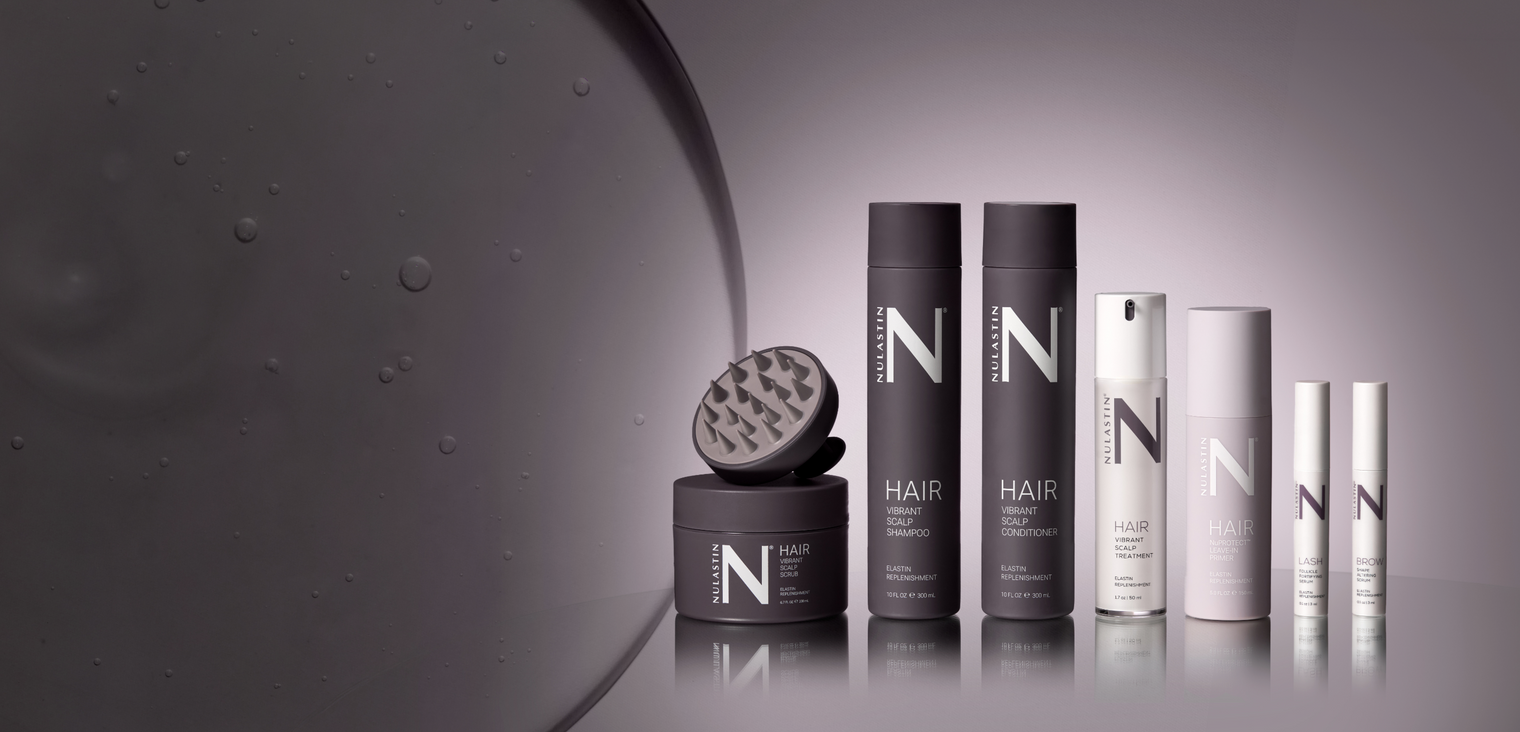 Front view of NULASTIN hair care product lineup on a reflective mauve background, showing bottles, jars, and an applicator tool