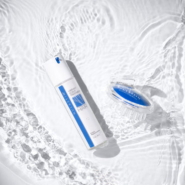 Top-down view of NULASTIN Vibrant Scalp Treatment bottle and clear blue scalp massager showing pump and bristles on rippling water background