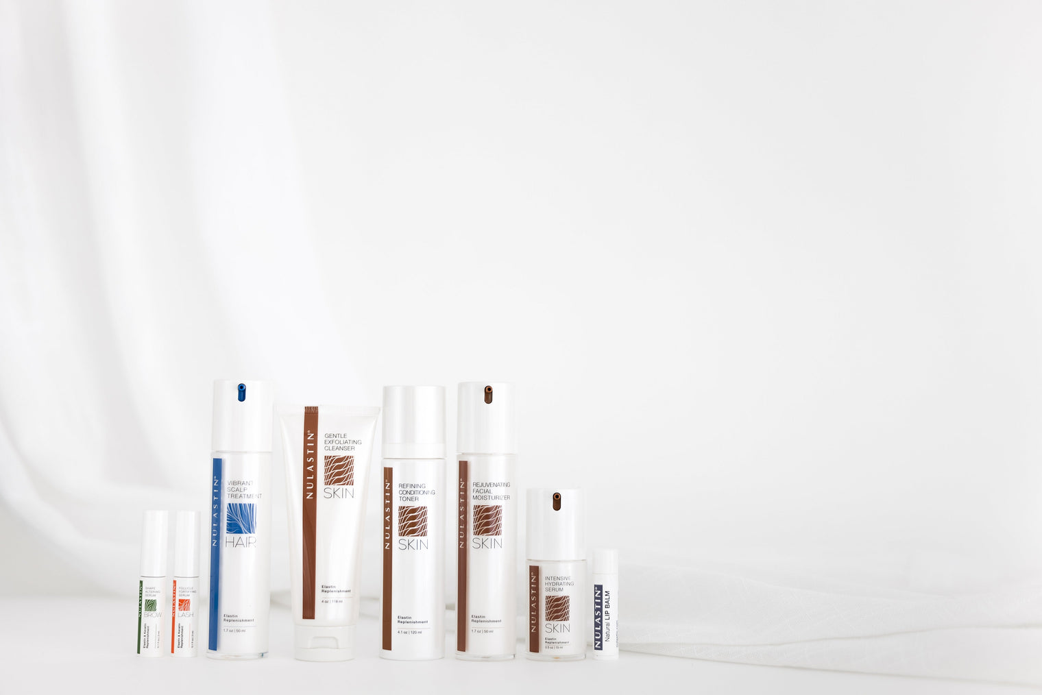 Front view of NULASTIN hair and skin product bottles and tubes lined up showcasing packaging and labels on a white draped background