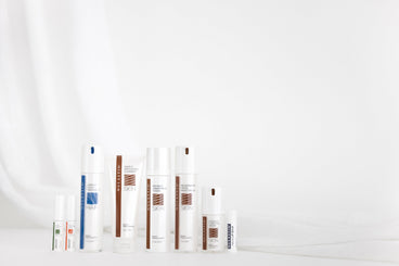 Front view of NULASTIN hair and skin product bottles and tubes lined up showcasing packaging and labels on a white draped background
