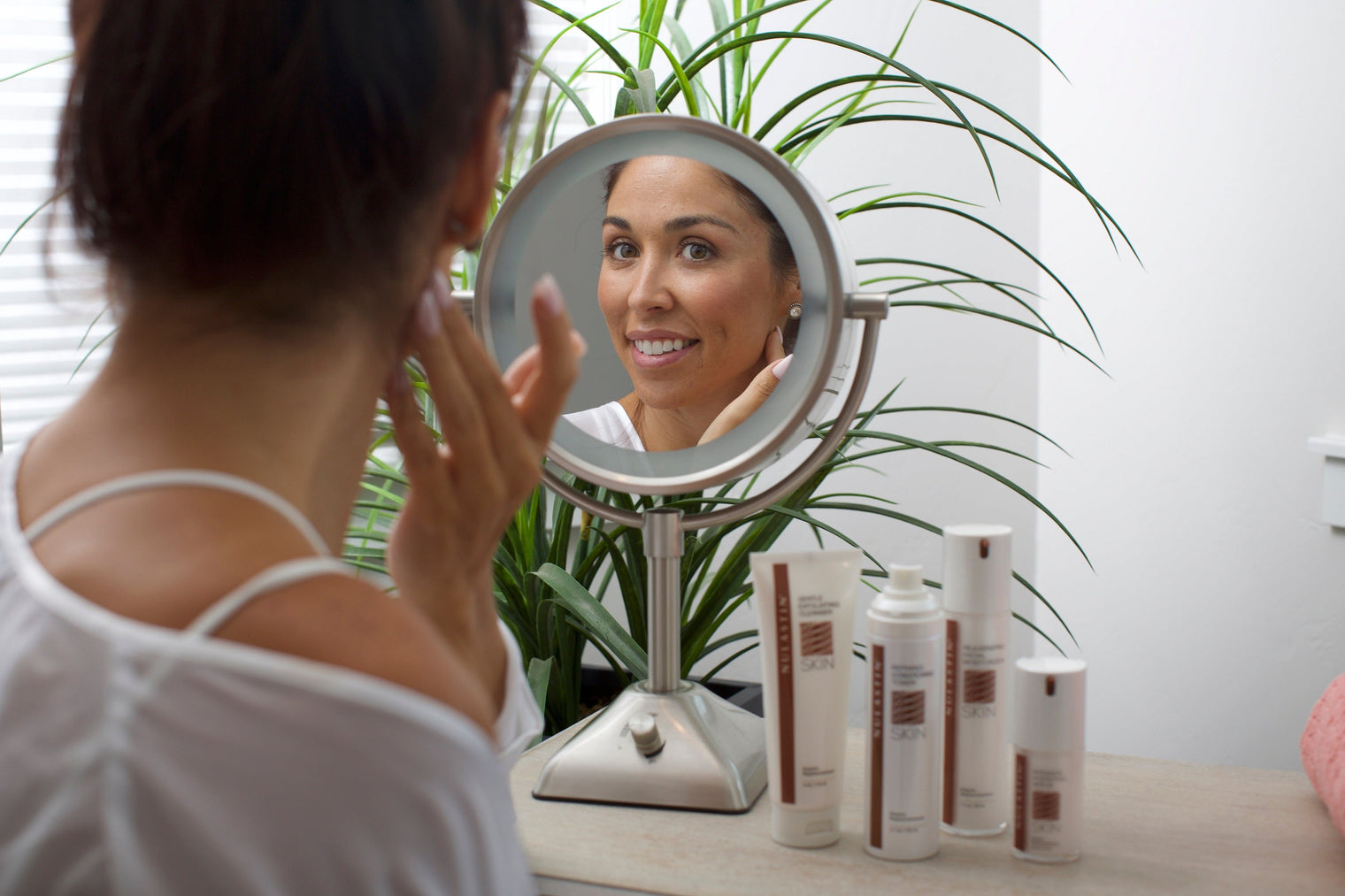Woman checking her face in a round vanity mirror while applying skincare, NULASTIN bottles on the vanity and a potted plant behind