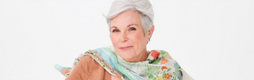 Smiling mature woman with short gray hair wearing a floral scarf and tan top against a white background