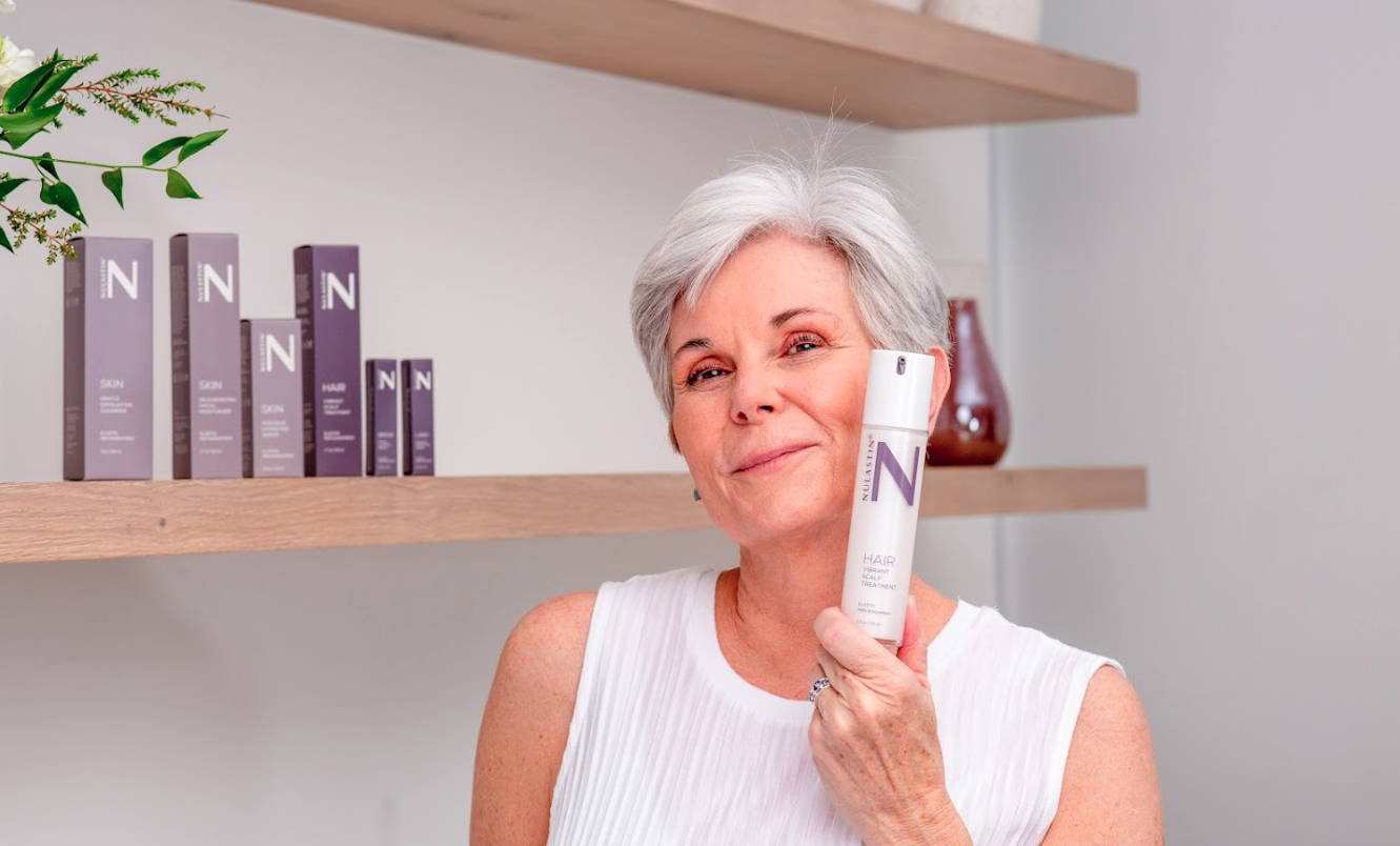 Front view of NULASTIN Hair Scalp Treatment bottle held by a smiling mature woman, product boxes on wooden shelf in background