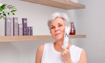 Front view of NULASTIN Hair Scalp Treatment bottle held by a smiling mature woman, product boxes on wooden shelf in background