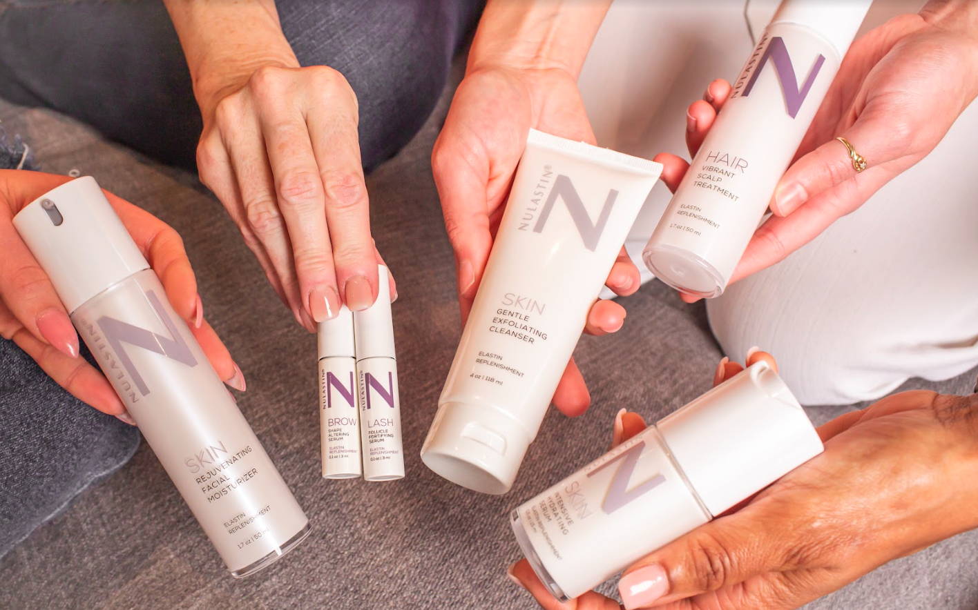 Top-down of NULASTIN products: cleanser tube, moisturizer, scalp treatment, and brow and lash serums on gray fabric
