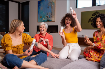Four women laughing and sharing NULASTIN skincare bottles while seated on a living-room sofa