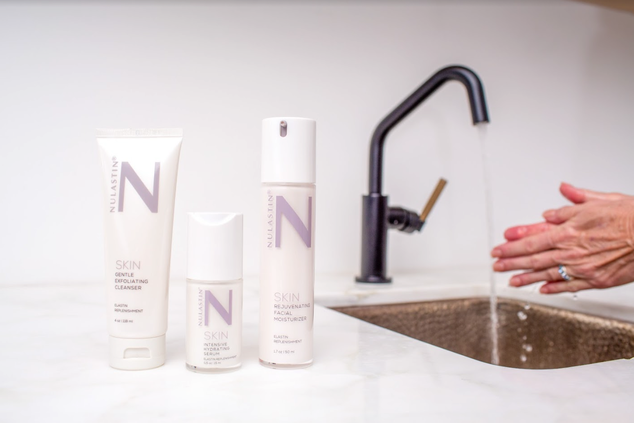 Three NULASTIN SKIN products — cleanser, hydrating serum, and facial moisturizer — on a marble counter with hands washing under a running faucet