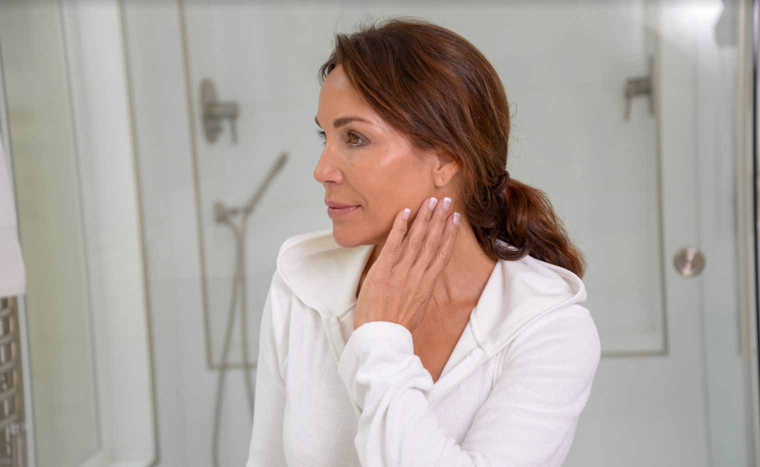 Woman in a white hoodie touching her jaw while looking in a bathroom mirror, bare-faced and relaxed