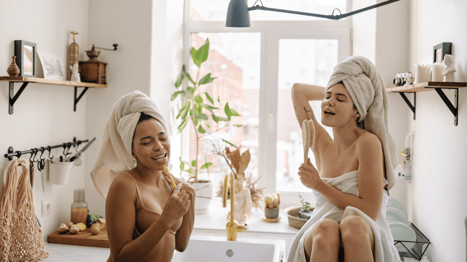 Two women with towels on their heads singing and playfully using a toothbrush and a body brush as microphones in a bright bathroom by a window