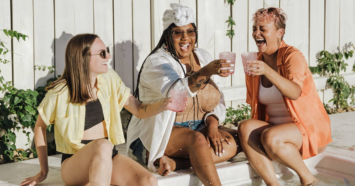 Three friends laughing and clinking pink glasses while sitting at the edge of a sunlit pool