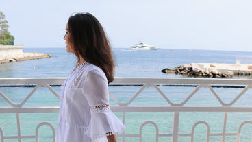 Woman in white lace cover-up standing on a seaside balcony overlooking turquoise water and a distant yacht