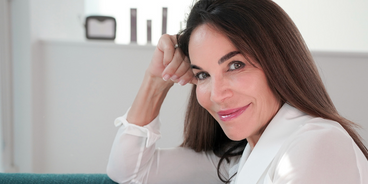 Smiling woman with long brown hair leaning on her hand in a bright, relaxed indoor setting