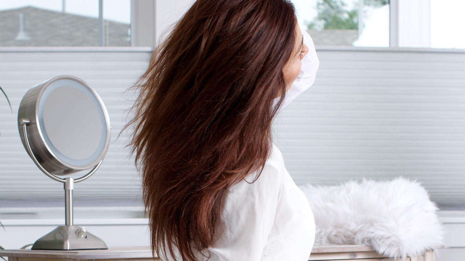 Woman with long brown hair flipping her hair at a vanity mirror in a bright room
