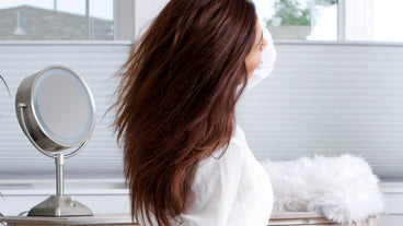 Woman with long brown hair flipping her hair at a vanity mirror in a bright room