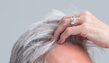Close-up of gray hair being lifted at the hairline by a hand wearing a silver ring against a neutral gray background