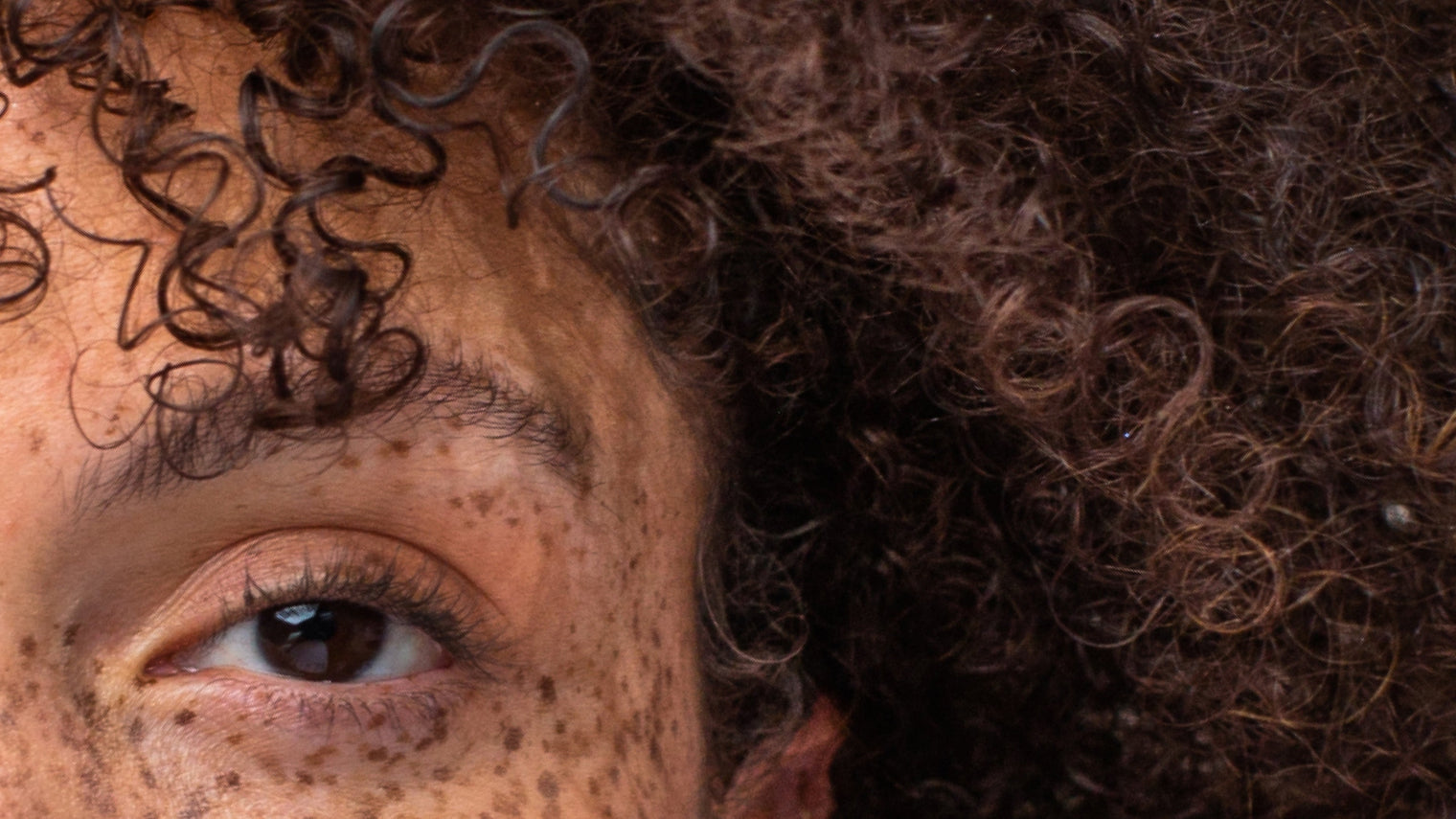 Close-up of left eye with freckles and dense brown curly hair showing hair texture