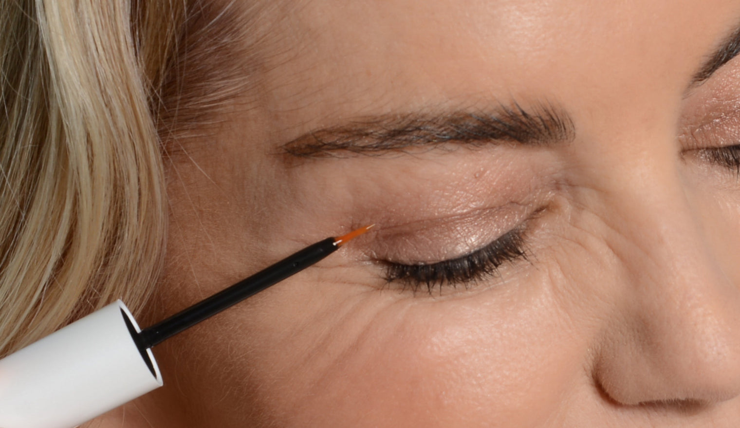 Woman applying lash serum to her upper lash line with a thin applicator brush, close-up of closed eye