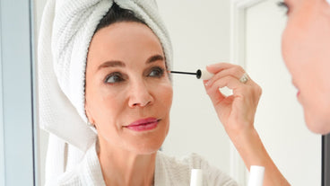 Woman in towel and robe applying a lash serum wand to her upper eyelashes while looking in a bathroom mirror