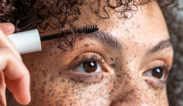 Close-up of a spoolie brush combing an eyebrow on freckled skin, showing short brow regrowth