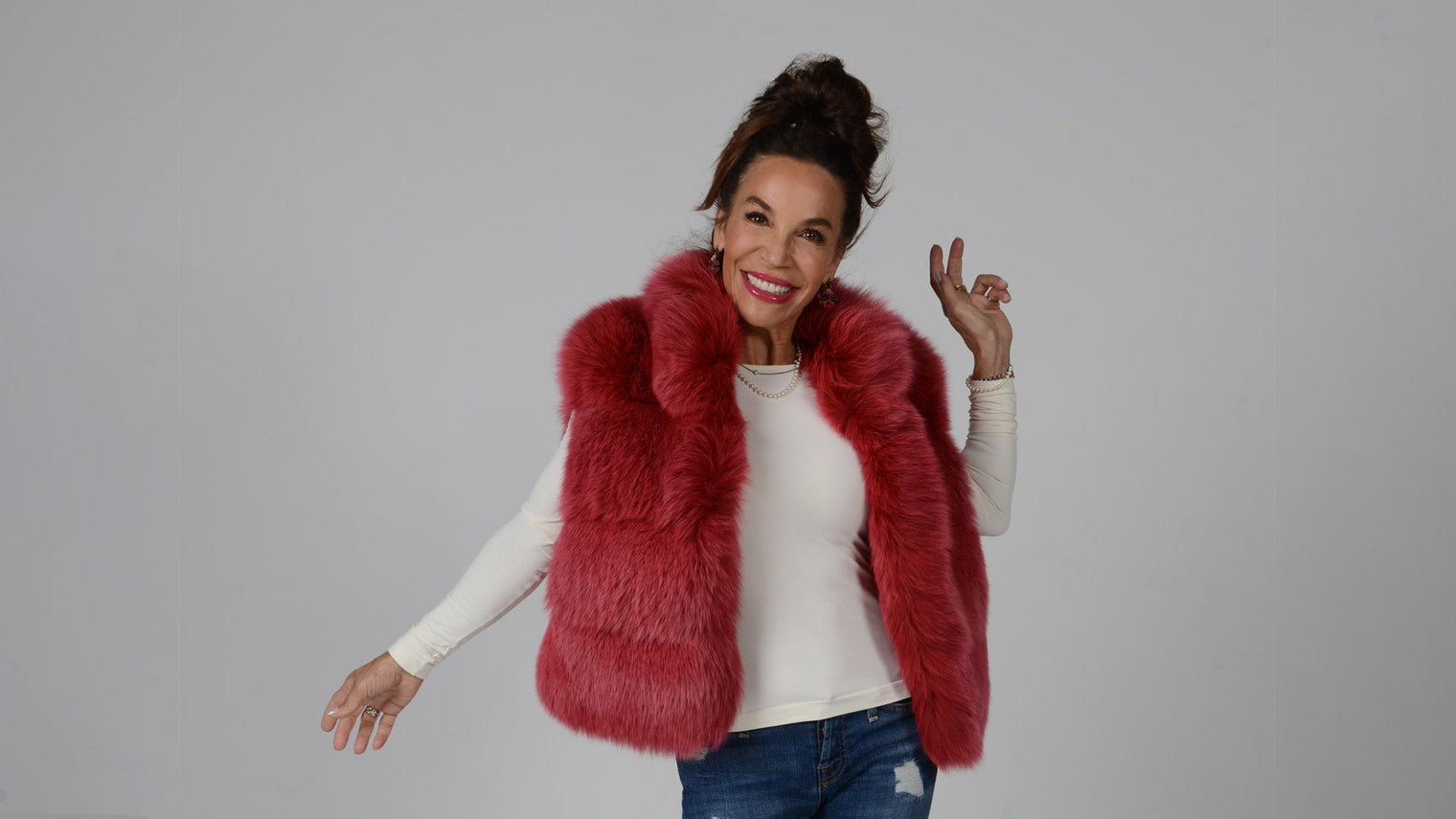 Smiling woman posing in a red faux-fur vest over a white long-sleeve top and distressed jeans against a light gray background
