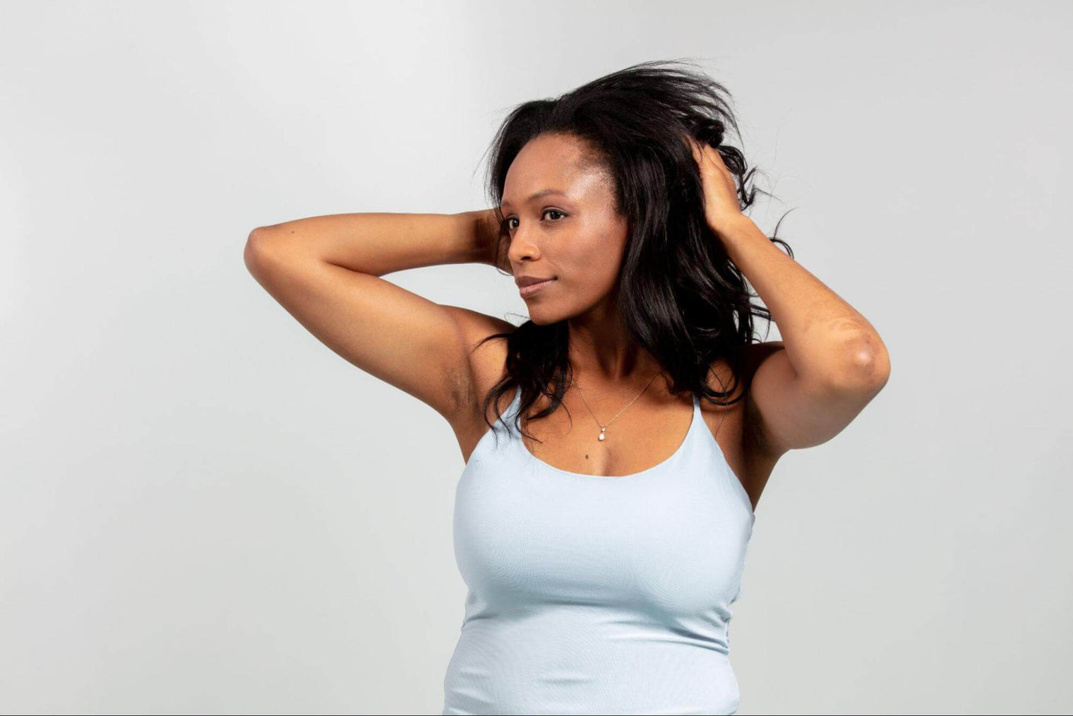 Woman running her hands through long dark hair, wearing a light blue fitted tank top against a light gray background