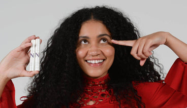 Woman smiling and pointing to her eye while holding two NULASTIN lash serums against a pale gray background