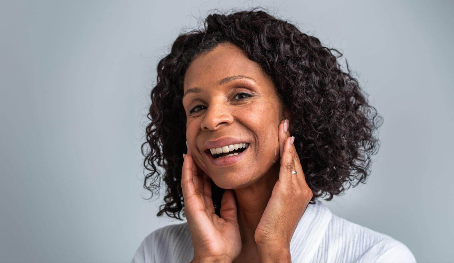 Middle-aged woman smiling and gently touching her face against a soft gray background
