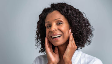 Middle-aged woman smiling and gently touching her face against a soft gray background