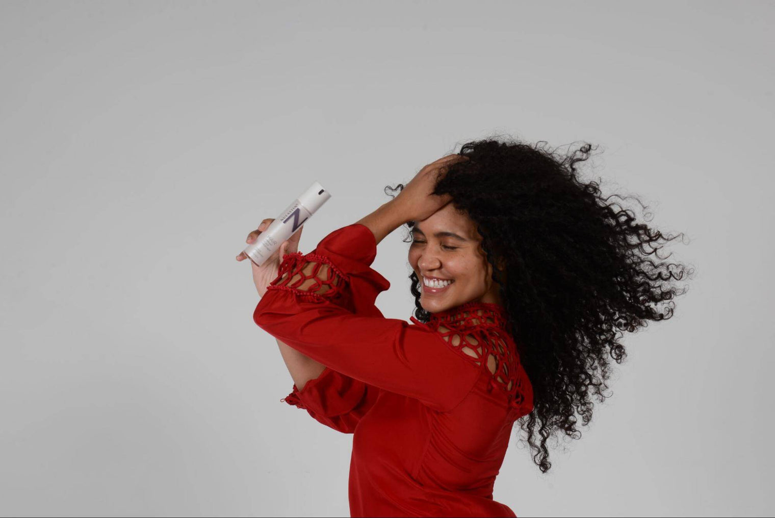 Woman with long curly hair smiling and holding a NULASTIN bottle near her scalp against a plain gray background