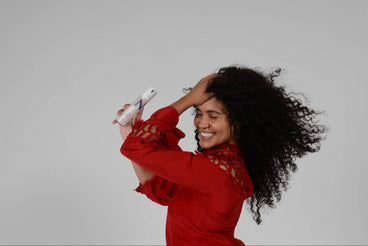 Woman with long curly hair smiling and holding a NULASTIN bottle near her scalp against a plain gray background
