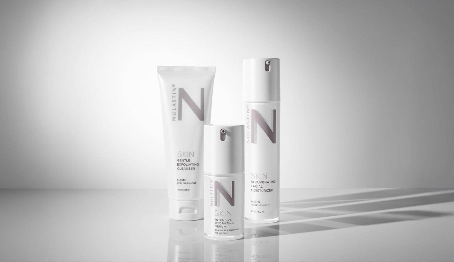 Front view of three NULASTIN SKIN products: cleanser, serum, moisturizer, white pump and tube on gray reflective background
