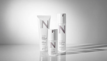 Front view of three NULASTIN SKIN products: cleanser, serum, moisturizer, white pump and tube on gray reflective background