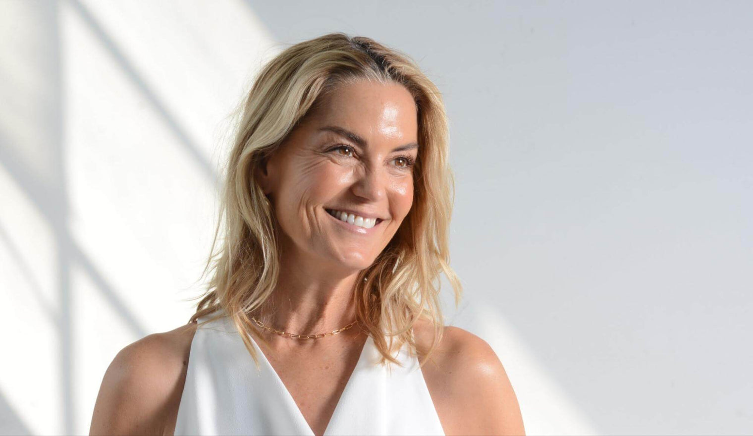 Smiling middle-aged woman with shoulder-length blonde hair and glowing skin looking off-camera against a bright neutral background