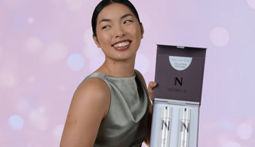 Woman holding open NULASTIN Solution Serums gift set with two serum bottles against a pink bokeh background