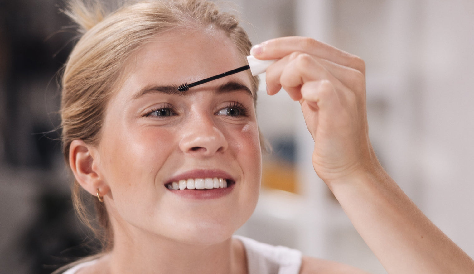 Young woman smiling while applying brow serum with a spoolie brush to her eyebrows in a bright bathroom