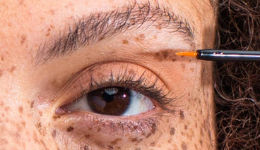 Close-up of a brown-eyed woman’s lashes and brow as a serum applicator brush touches the skin, showing natural freckles