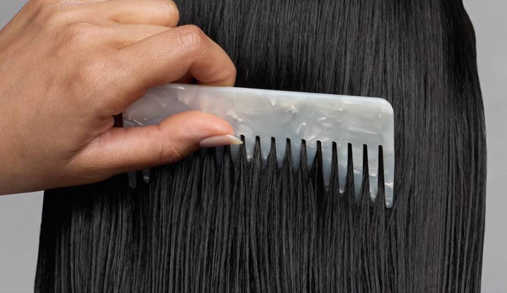 Hand combing long glossy black hair with a wide-tooth pearlescent comb against a neutral gray background