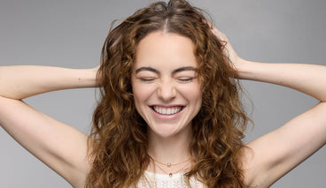 Young woman with curly brown hair smiling with eyes closed and hands behind her head against a gray background