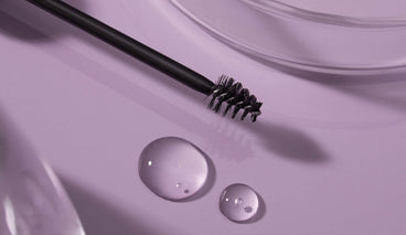 Close-up of a black spoolie brow brush with serum on the bristles and two serum droplets on a lavender surface