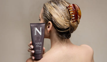 Front view of NULASTIN Vibrant Scalp Mask (200 mL) tube held against a woman’s damp hair, beige background