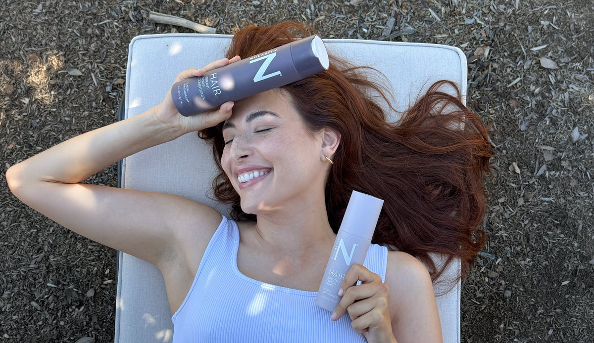 Smiling woman with long red hair lying outdoors on a cushion, holding two NULASTIN hair product bottles