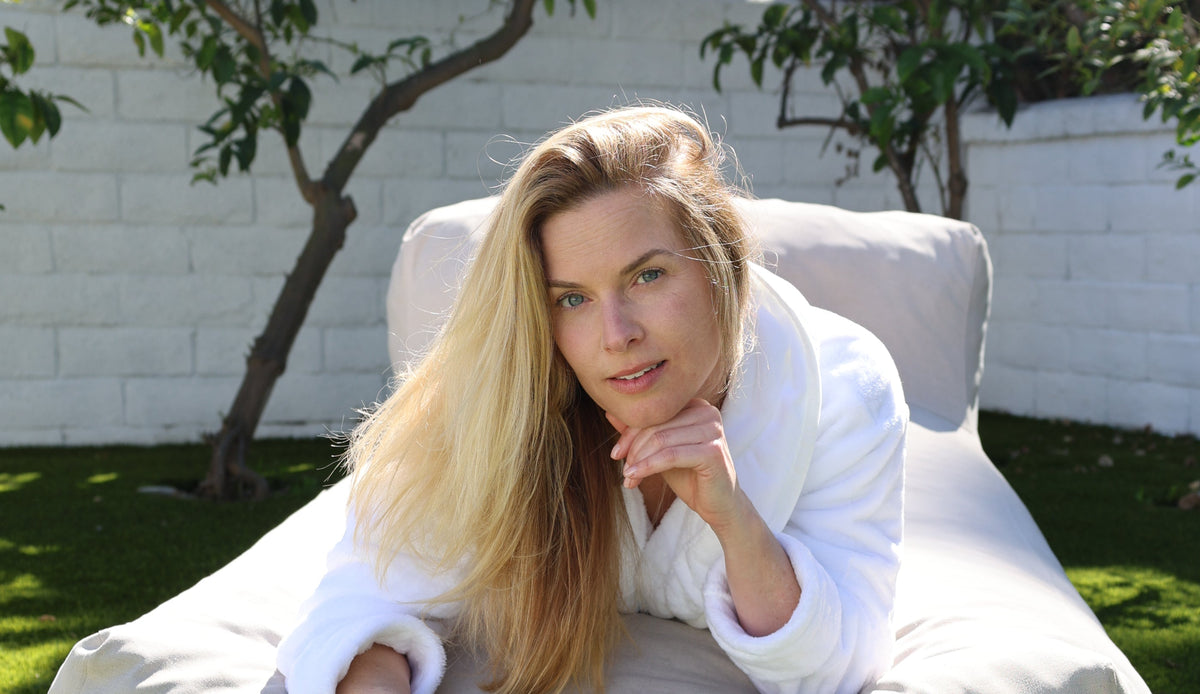Sunlit woman reclining on outdoor lounge chair in white robe, resting chin on hand in a backyard with a white brick wall