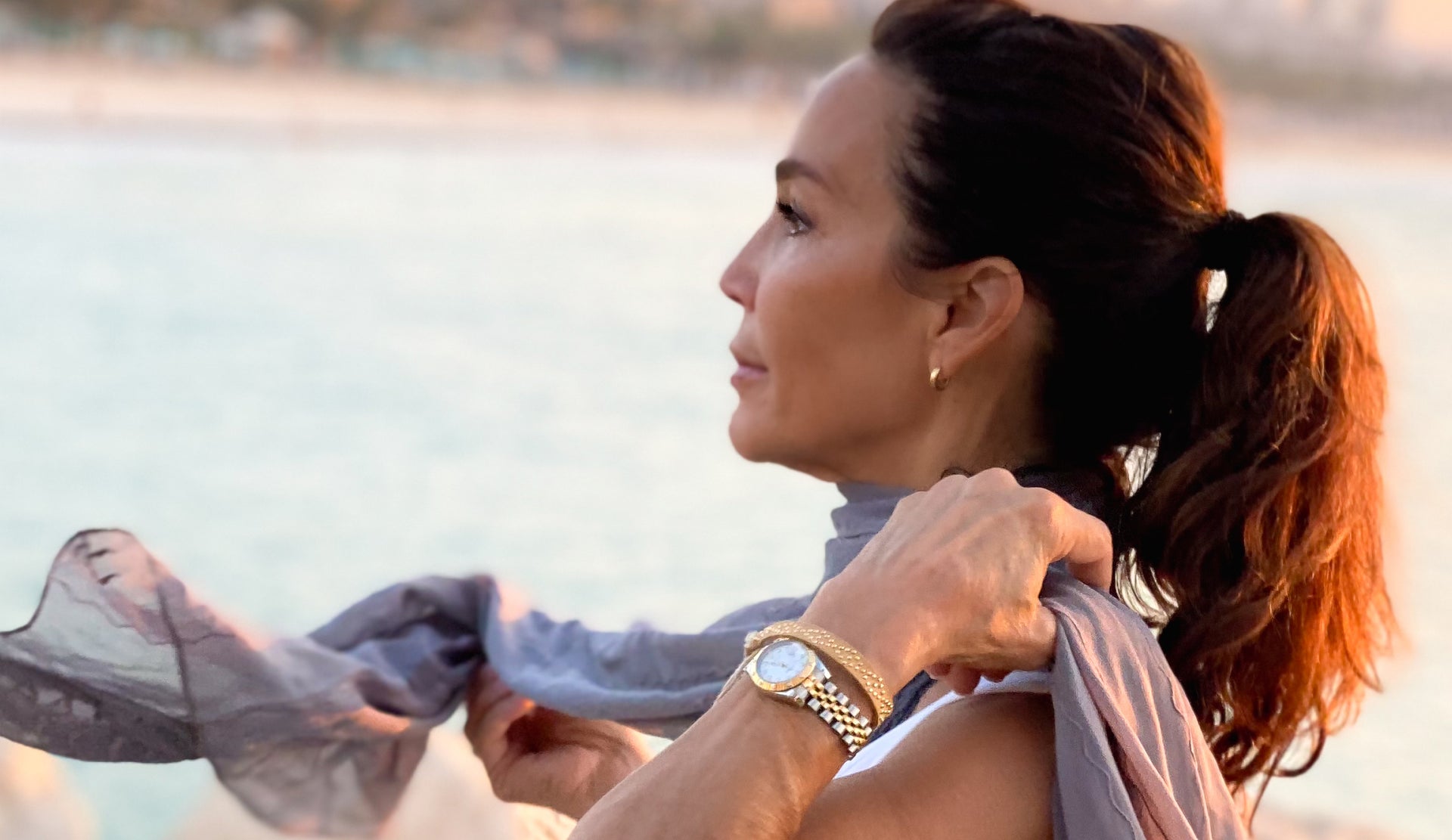 Beautiful brunette woman wearing chic gold jewelry adjusts her windblown scarf as she looks hopefully into the distance at golden hour