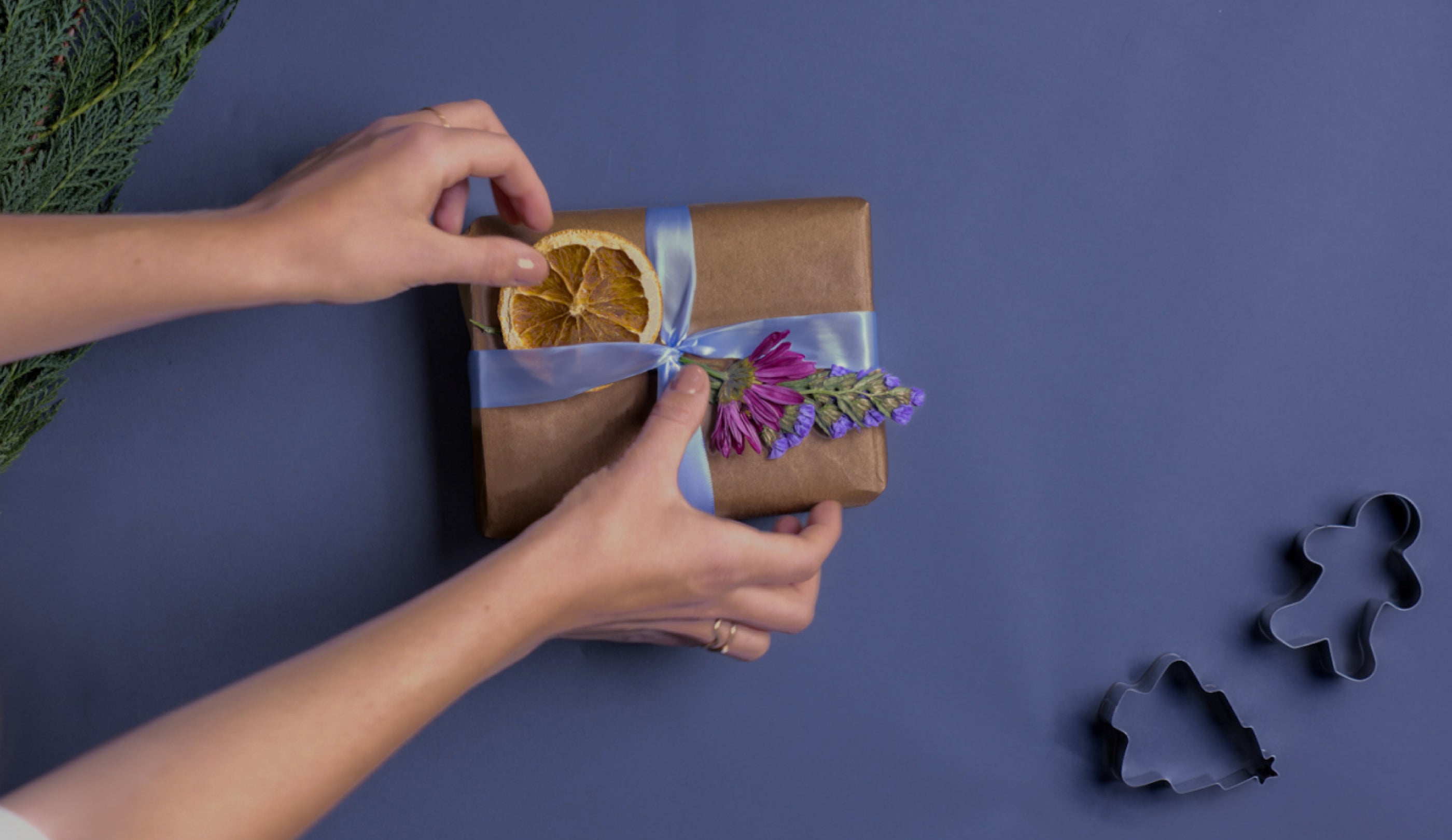 A woman’s forearms on a smooth surface with her hands are wrapping a gift with Kraft paper. She is using ribbon, a sprig of flowers, and a slice of dried orange to decorate package.